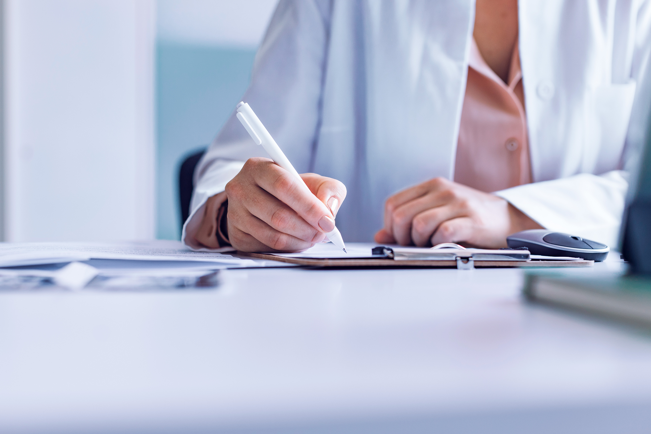 A doctor in a white coat takes notes on a clipboard while consulting with a patient in an exam room